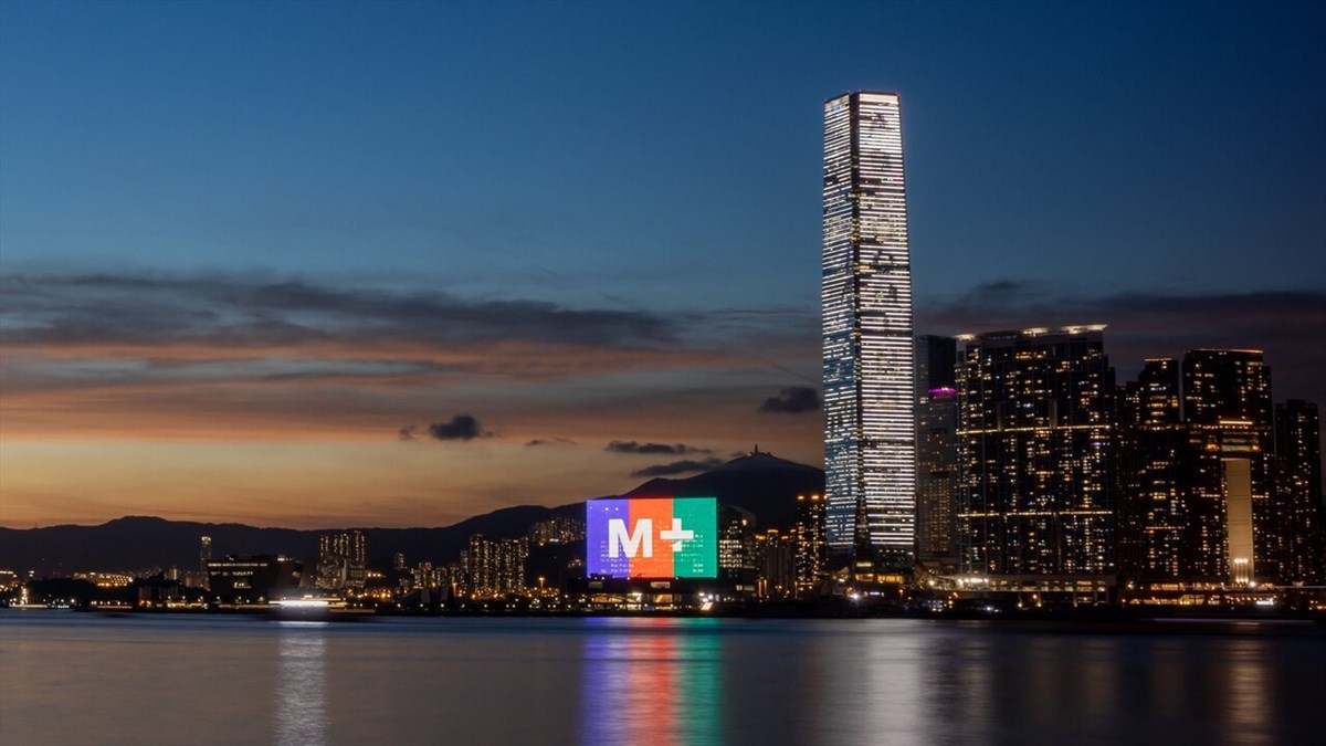 M+ Facade Biggest LED Screen Project LED Display Manufacturer in China EagerLED Dusk skyline of a harbor city with a tall illuminated skyscraper and a colorful M+ billboard reflected in calm water