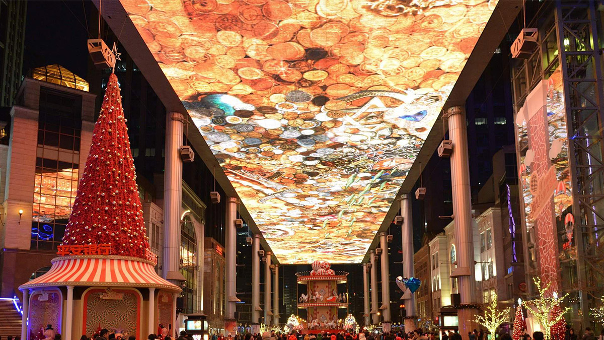 The Place Sky Screen Biggest LED Screen Project LED Display Manufacturer in China EagerLED Festive nighttime street scene at a mall with a ceiling covered in coin shaped art a tall red Christmas tree on the left and shoppers below