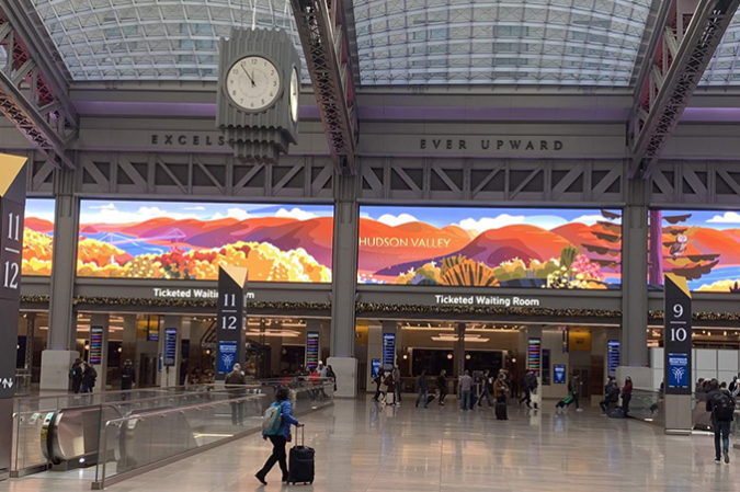 Cos'è uno schermo pubblicitario a LED? -1 L'affollato atrio della stazione ferroviaria è caratterizzato da un grande murale colorato, un orologio centrale e la segnaletica per la sala d'attesa riservata ai possessori di biglietto; i viaggiatori con bagagli si spostano tra i binari 11 e 12 in una sala con tetto in vetro.