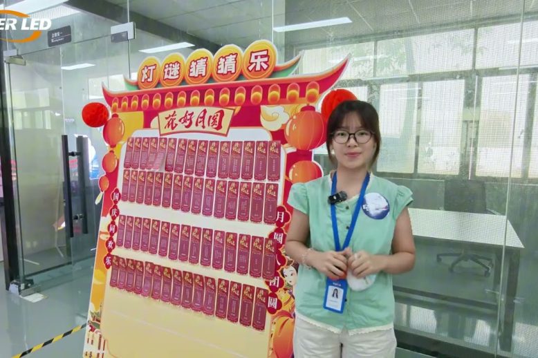 descargar Una mujer con gafas y un cordón para el cuello está de pie junto a un gran panel de exhibición colorido lleno de tarjetas rojas y faroles festivos en un entorno de oficina con paredes de cristal.